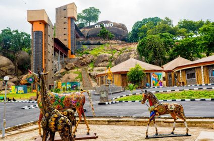 The main courtyard of Olumo Rock Tourist Centre featuring colorful animal sculptures, a central statue, thatched-roof buildings, and the glass elevator towers leading up to the historic rock.